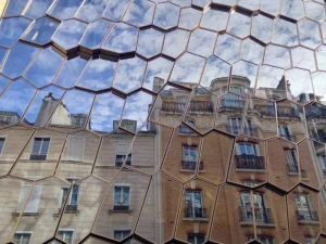 Cette photographie met en scène deux bâtiments aux architectures contrastées. Le sujet principal est la juxtaposition entre un immeuble traditionnel, aux lignes classiques et aux tons beiges, et une structure moderne recouverte de miroirs hexagonaux déformants.Le cadrage est serré, mettant en valeur les reflets et les angles géométriques. La lumière est naturelle, éclatante, créant des jeux d'ombres et de reflets. Les couleurs dominantes sont les tons neutres (beige, gris) et les teintes bleutées du ciel.Le style est urbain et contemporain, évoquant une ambiance futuriste et artistique.
