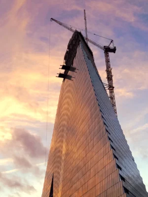 Cette photographie montre un gratte-ciel en construction, sujet principal. La composition met en valeur la pointe de l'immeuble avec une perspective ascendante. Le cadrage serré isole la structure, avec une grue en haut.La lumière dorée du coucher de soleil crée un contraste saisissant. Les couleurs dominantes sont les tons chauds orangés et roses, contrastant avec le gris métallique du bâtiment. Le style est réaliste et documentaire. L’ambiance est à la fois dynamique et contemplative, évoquant la puissance architecturale et la fin d'une journée.