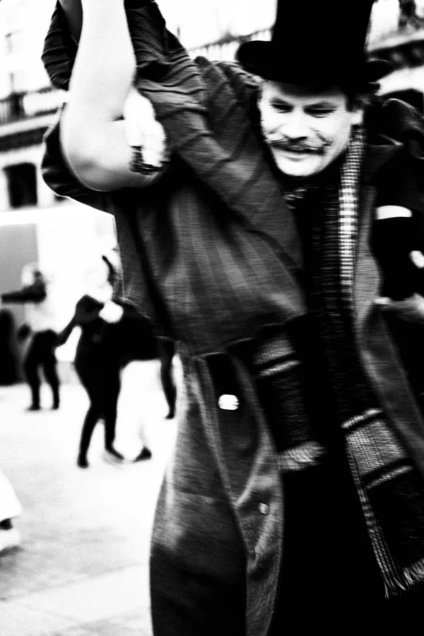 Thierry · 03/01/2026 · 2560 x 3840 Cette photographie en noir et blanc met en scène un homme comme sujet principal, vêtu d’un long manteau et d’un chapeau, levant un bras avec un geste théâtral. La composition est centrée sur lui, avec un cadrage serré qui isole sa silhouette. L’arrière-plan flou montre des silhouettes indistinctes, suggérant une rue animée.La lumière semble naturelle, contrastée, accentuant les ombres et les détails du visage et des vêtements. L’ambiance est dramatique et mystérieuse, renforcée par le monochrome. Le style rappelle le cinéma classique ou les photographies documentaires des époques passées.