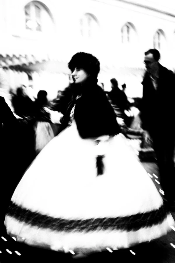 Thierry · 03/01/2026 · 2560 x 3840 Cette photographie en noir et blanc met en scène une femme vêtue d'une robe à crinoline, sujet principal du cliché. La composition montre une ambiance festive ou historique, avec des silhouettes floues en arrière-plan. Le cadrage serré sur la femme accentue son costume volumineux et son expression. La lumière semble douce, créant des contrastes subtils. Les tons dominants sont les gris et noirs, typiques du monochrome. Le style évoque une époque ancienne, peut-être le XIXe siècle. L'ambiance est à la fois élégante et nostalgique, avec une touche de mouvement suggéré par le flou des autres personnages.