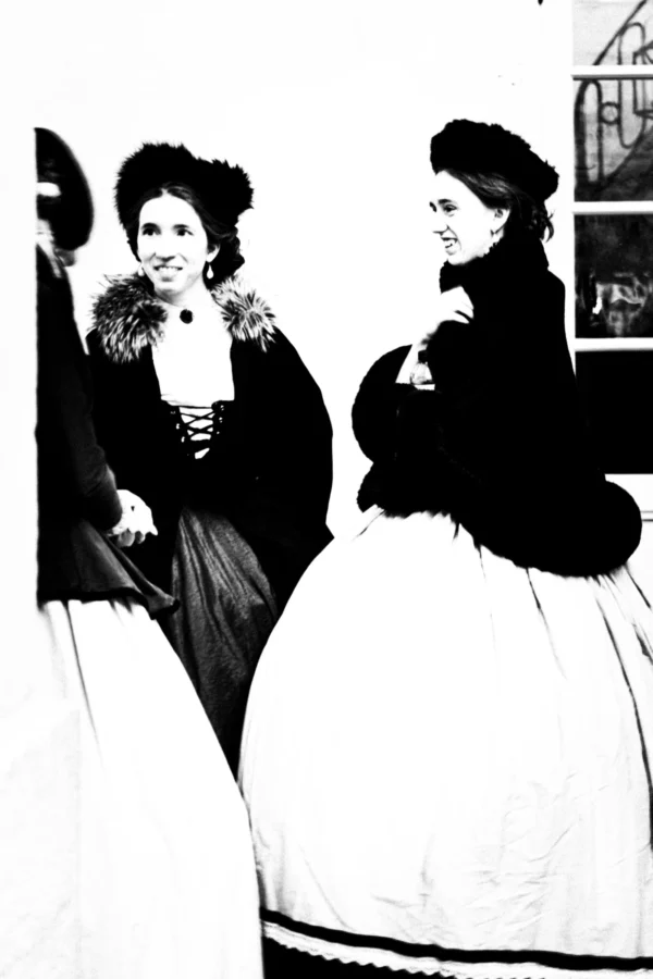 Thierry · 03/01/2026 · 2560 x 3840 Cette photographie en noir et blanc montre deux femmes vêtues de robes élégantes du XIXe siècle, probablement des costumes d'époque. Les sujets principaux sont les deux femmes, assises et souriantes, avec des détails comme les chapeaux ornés et les manteaux à volants.La composition est symétrique et serrée, avec un cadrage rapproché. La lumière est douce et semble naturelle, éclairant les visages et les détails des tenues. Les couleurs dominantes sont les tons de gris, typiques du noir et blanc, accentuant les contrastes.Le style évoque une ambiance historique et théâtrale, renforcée par les accessoires et les poses. L'atmosphère est chaleureuse et nostalgique.