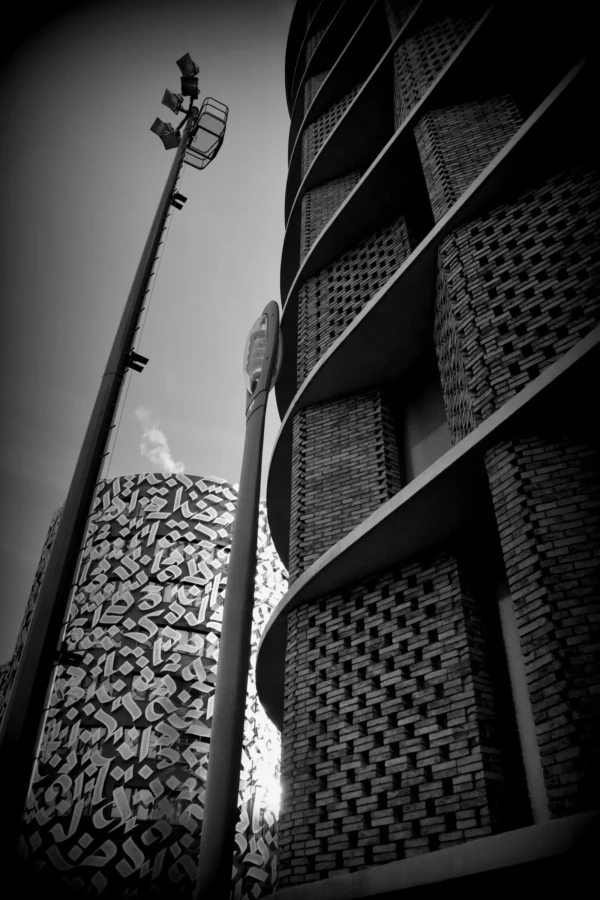 Vincent · 17/01/2026 · 3500 x 5250 Cette photographie en noir et blanc met en scène une architecture moderne et géométrique. Le sujet principal est un bâtiment aux formes anguleuses et aux façades ajourées, évoquant une structure contemporaine. La composition joue sur les lignes verticales et horizontales, renforçant la sensation de hauteur et de modernité.Le cadrage serré accentue les détails architecturaux et les jeux d'ombres. La lumière, contrastée, crée des effets dramatiques avec des zones très claires et d'autres profondément sombres. L'absence de couleurs et les jeux de lumière renforcent l'ambiance urbaine et minimaliste, presque abstraite. Le style est épuré et artistique.