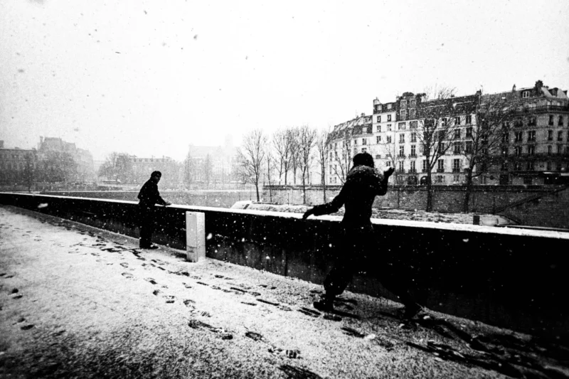 Thierry · 05/01/2026 · 3840 x 2560 Cette photographie en noir et blanc capture une scène hivernale urbaine. Le sujet principal est un homme en manteau, marchant sur un pont enneigé, tandis qu’une autre personne, partiellement visible, semble l’observer.La composition est dynamique, avec une perspective légèrement basse. Le cadrage encadre le pont et les bâtiments en arrière-plan, créant une profondeur. La lumière est tamisée, typique d’un jour froid et brumeux, renforçant l’ambiance mélancolique.Les tons dominants sont les gris et noirs, accentués par la neige. Le style, sobre et réaliste, évoque une époque ancienne, renforçant une atmosphère nostalgique et contemplative.