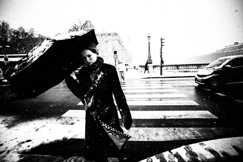 Thierry · 05/01/2026 · 3840 x 2560 Cette photographie en noir et blanc met en scène une personne tenant un parapluie, sujet principal partiellement masqué. La composition joue sur les contrastes et les formes géométriques, avec des lignes de trottoir et des véhicules en arrière-plan.Le cadrage est serré, centré sur le sujet, créant une ambiance intime. La lumière semble diffuse, typique d’un jour pluvieux, renforçant l’atmosphère mélancolique. Les tons dominants sont les gris et noirs, accentuant le côté dramatique et poétique. Le style rappelle le photojournalisme classique ou une approche artistique inspirée du réalisme urbain.