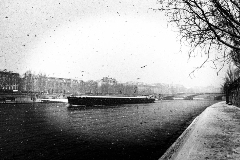 Thierry · 05/01/2026 · 3840 x 2560 Cette photographie en noir et blanc montre un bateau de chargement navigant sur une rivière. Le sujet principal est le bateau, centré dans le cadre.La composition est équilibrée, avec une rive bordée de bâtiments et d’arbres dénudés à gauche, et une berge en pierre à droite. Le cadrage est large, incluant une partie du paysage urbain en arrière-plan.La lumière est tamisée, créant une ambiance nostalgique et légèrement brumeuse. Les tons dominants sont les gris et noirs, typiques du style sépia. L’ambiance est calme et mélancolique, évoquant une époque passée.