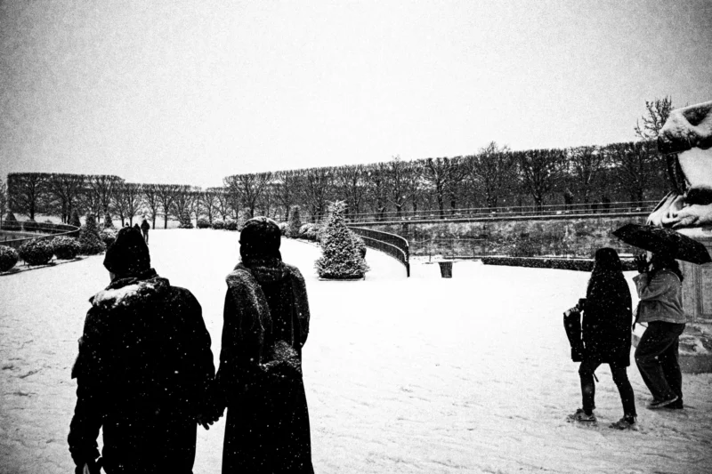 Thierry · 05/01/2026 · 3840 x 2560 Cette photographie en noir et blanc capture un groupe de personnes enneigées, dont le sujet principal semble être trois silhouettes au premier plan. La composition met en valeur un espace ouvert et une architecture lointaine, probablement un monument. Le cadrage est large, incluant un ciel couvert et une étendue neigeuse. La lumière est douce et diffuse, typique d’un jour hivernal. Les couleurs dominantes, bien que monochromes, évoquent des tons de gris et de blanc. Le style est documentaire et sobre, créant une ambiance calme et mélancolique, renforcée par la solitude des figures dans un paysage enneigé.