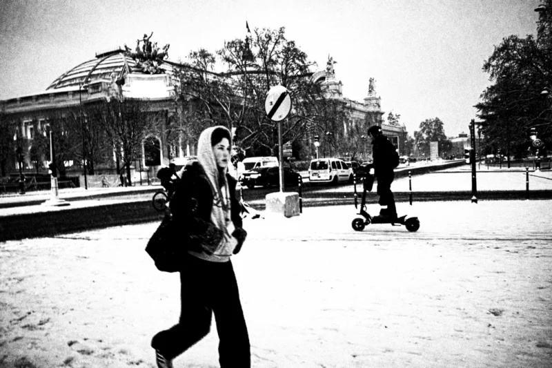 Thierry · 05/01/2026 · 3840 x 2560 Cette photographie en noir et blanc met en scène une personne marchant avec un sac à dos, comme sujet principal. La composition montre aussi un individu sur un scooter électrique à l’arrière-plan.Le cadrage est serré sur la personne en mouvement, avec une profondeur de champ qui inclut l’environnement urbain. La lumière est douce, typique d’un jour couvert ou d’un temps froid, créant des contrastes modérés.L’ambiance est calme et légèrement mélancolique, renforcée par le monochrome. Les tons dominants varient du gris clair au noir, accentuant les textures et les formes architecturales. Le style rappelle un documentaire urbain intemporel.