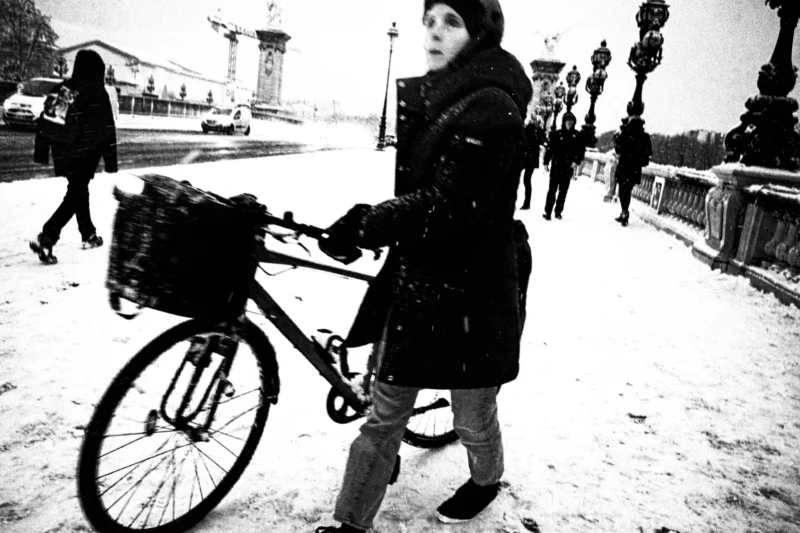 Thierry · 05/01/2026 · 3840 x 2560 Cette photographie en noir et blanc met en scène une jeune femme comme sujet principal, vêtue d’un long manteau, tenant un vélo chargé d’un panier. La composition est simple et centrée sur elle, avec un cadrage serré qui isole le sujet de l’arrière-plan.La lumière, douce et diffuse, crée des contrastes modérés. L’ambiance est hivernale et mélancolique, renforcée par la neige au sol et les vêtements épais. Le style est documentaire et intime, évoquant une scène de vie quotidienne dans un cadre urbain. Les tons dominants, en niveaux de gris, accentuent la froideur et la sérénité du moment.