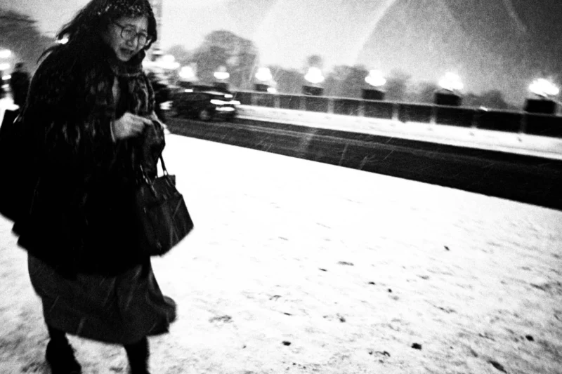Thierry · 05/01/2026 · 3840 x 2560 Cette photographie en noir et blanc met en scène une femme comme sujet principal, vêtue d’un manteau épais et tenant un sac. Elle semble marcher dans un environnement enneigé et urbain.La composition est simple et centrée, avec un cadrage serré sur la personne, isolée sur un pont. La lumière est faible, créant une ambiance sombre et contrastée. Les couleurs dominantes sont les nuances de gris, renforçant l’atmosphère froide et mélancolique.Le style est documentaire et intemporel, évoquant une scène réaliste et poétique, imprégnée de solitude et de sérénité hivernale.