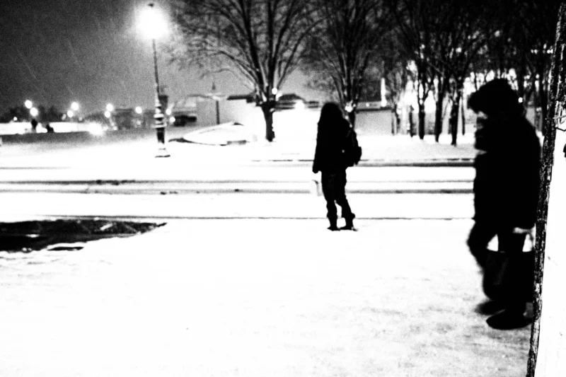 Thierry · 05/01/2026 · 3840 x 2560 Cette photographie en noir et blanc capture deux personnes marchant dans un parc enneigé. Le sujet principal semble être l’un des individus, légèrement flou, tandis que l’autre est plus net, créant une dynamique de profondeur.La composition repose sur une ligne de perspective menant vers l’arrière-plan, avec des arbres et des lumières en toile de fond. Le cadrage est serré, mettant l’accent sur les silhouettes et l’atmosphère.Les tons dominants sont les gris et noirs, accentués par la lumière artificielle tamisée, renforçant une ambiance froide et mélancolique. Le style est documentaire et poétique, évoquant solitude et sérénité hivernale.