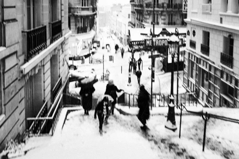 Thierry · 07/01/2026 · 3840 x 2560 Cette photographie en noir et blanc capture une scène hivernale urbaine. Le sujet principal est un groupe de personnes descendant une rue enneigée en pente, dont un homme glissant avec un sac. La composition est dynamique, avec une perspective aérienne montrant des bâtiments de chaque côté et des passants en mouvement.Le cadrage inclut des détails architecturaux et des panneaux de signalisation. La lumière est douce, typique d’un jour hivernal couvert. Les tons dominants sont les gris et noirs, accentués par la neige. Le style est documentaire, évoquant une ambiance froide et vivante, presque théâtrale, reflétant la vie quotidienne dans un environnement rigoureux.