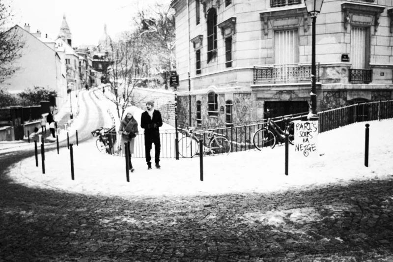 Thierry · 07/01/2026 · 3840 x 2560 Cette photographie en noir et blanc capture deux hommes marchant côte à côte sur un trottoir enneigé. Le sujet principal est leur silhouette, entourée d’une ambiance urbaine hivernale.La composition met en valeur la perspective avec des bâtiments et des balustrades en arrière-plan. Le cadrage est serré, centré sur les personnages et leur environnement immédiat. La lumière est douce, créant des contrastes subtils entre ombres et clartés. Les couleurs dominantes, bien que monochromes, évoquent des tons gris et blancs, renforçant l’atmosphère froide et nostalgique.Le style est documentaire, réaliste, avec une touche mélancolique. L’ambiance est calme et introspective, reflétant la sérénité d’un hiver urbain.