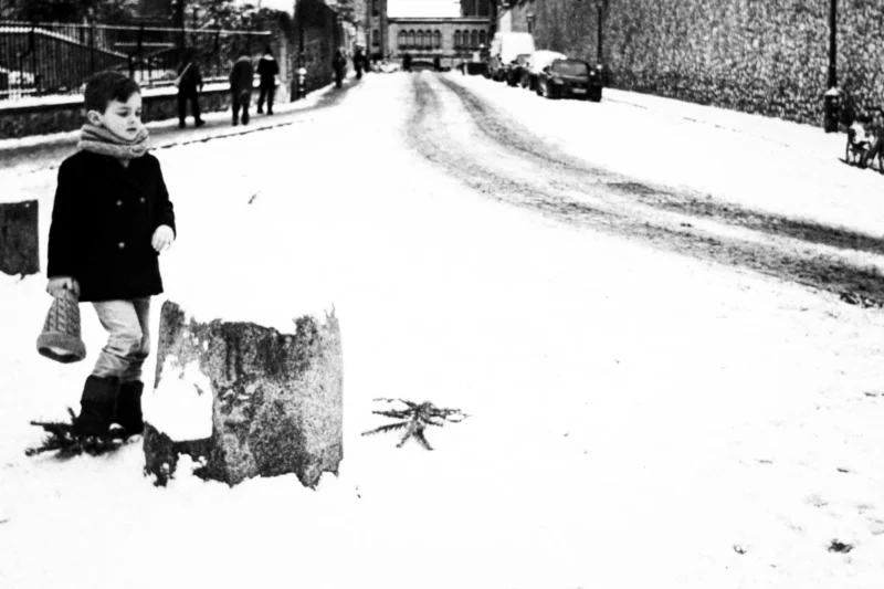 Thierry · 07/01/2026 · 3840 x 2560 Cette photographie en noir et blanc capture une scène hivernale urbaine. Le sujet principal est une personne, probablement une enfant, tenant des objets et marchant dans la neige. La composition montre une rue enneigée avec des traces de pas et des déchets, évoquant une ambiance modeste.Le cadrage est serré sur la personne, avec une profondeur de champ qui met en valeur les détails du sol. La lumière semble naturelle, probablement diffuse, créant des contrastes doux. Les couleurs dominantes sont les tons de gris et de noir, renforçant l’aspect froid et mélancolique. Le style est documentaire, évoquant une époque passée avec une ambiance sobre et réaliste.