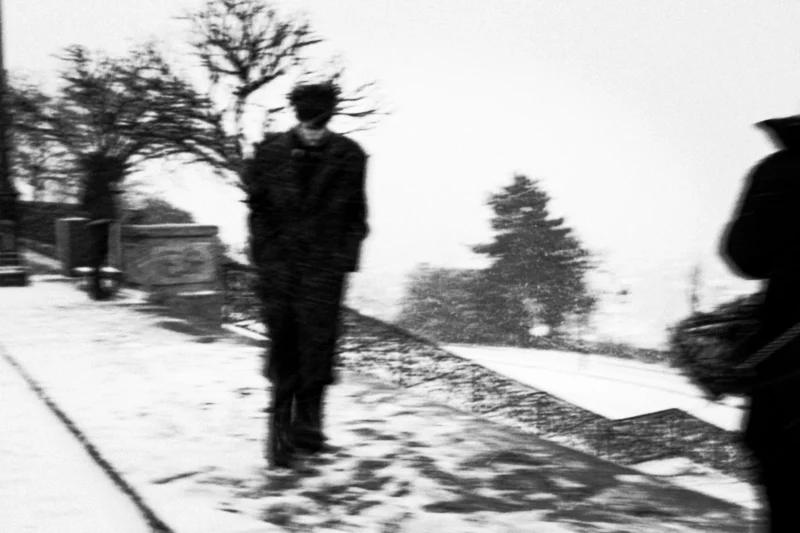 Thierry · 07/01/2026 · 3840 x 2560 Cette photographie en noir et blanc met en scène un homme comme sujet principal, marchant seul sur un trottoir enneigé. La composition est simple, avec une perspective légèrement basse et un cadrage serré sur le sujet, qui semble isolé.La lumière est diffuse, typique d’un jour hivernal, créant des contrastes doux. Les couleurs dominantes sont les nuances de gris, renforçant l’atmosphère froide et monochrome.Le style est documentaire, presque vintage, évoquant une ambiance mélancolique et introspective, renforcée par le flou et la neige qui enveloppe la scène.