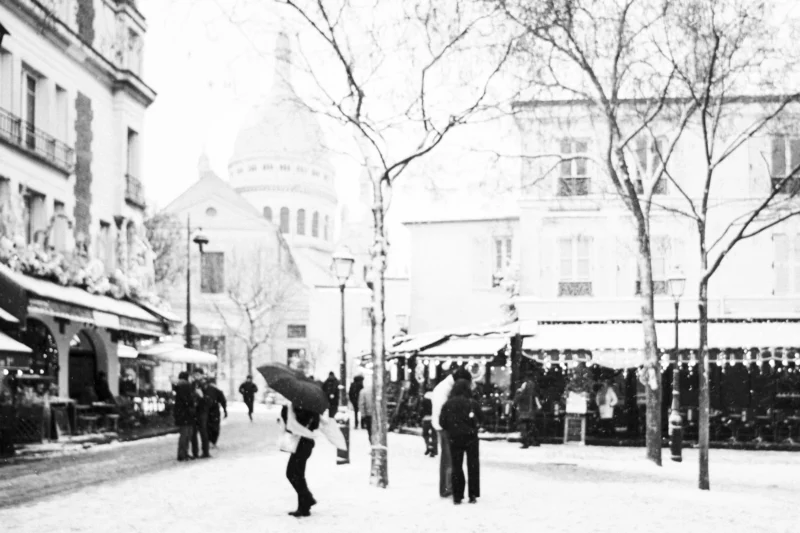 Thierry · 07/01/2026 · 3840 x 2560 Cette photographie en noir et blanc capture une scène urbaine animée. Le sujet principal semble être un homme en mouvement au premier plan, ajoutant une dynamique à l'image. La composition met en valeur une avenue bordée de cafés et d'arbres dénudés, avec des passants en arrière-plan.Le cadrage est large, englobant une partie de la rue et des bâtiments. La lumière est douce, typique d'un jour couvert ou d'un hiver ensoleillé, créant des contrastes subtils. Les couleurs dominantes, bien que monochromes, varient en tons de gris et noirs, renforçant l'atmosphère.Le style est documentaire et intemporel, évoquant une ambiance calme et mélancolique, presque nostalgique.