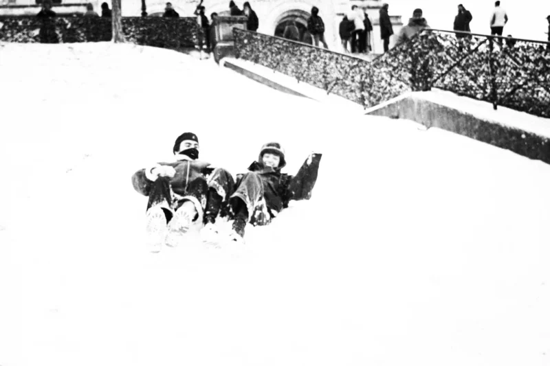 Thierry · 07/01/2026 · 3840 x 2560 Cette photographie en noir et blanc montre deux enfants assis sur une pente enneigée, semblant glisser ou s’amuser. Le sujet principal est leur action ludique et insouciante.La composition met en valeur les enfants au premier plan, avec un cadrage serré. En arrière-plan, on distingue des spectateurs et une structure métallique, ajoutant de la profondeur. La lumière semble naturelle, probablement en journée, créant des contrastes nets. Les tons dominants sont les gris et noirs, typiques du noir et blanc, renforçant l’ambiance nostalgique et hivernale. Le style est documentaire et spontané, évoquant une scène authentique et joyeuse.