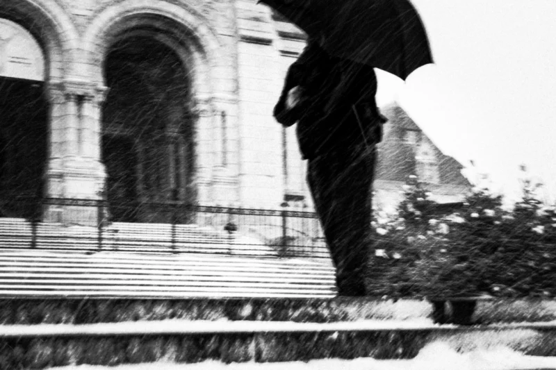Thierry · 07/01/2026 · 3840 x 2560 Cette photographie en noir et blanc met en scène une personne tenant un parapluie, marchant sous la neige. Le sujet principal est flou, suggérant un mouvement rapide.La composition est dynamique, avec un cadrage serré sur les jambes et le parapluie, tandis que l'arrière-plan, une architecture classique, est légèrement flouté. La lumière est douce et diffuse, typique d’un jour neigeux.Les tons dominants sont les gris et noirs, renforçant l’ambiance froide et mélancolique. Le style est documentaire et poétique, évoquant une atmosphère contemplative et introspective.