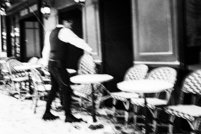 Thierry · 07/01/2026 · 3840 x 2560 Cette photographie en noir et blanc met en scène un homme en mouvement, sujet principal, marchant devant des tables et chaises de café empilées. La composition est dynamique, avec un cadrage serré sur le sujet en bas à gauche, créant un effet de profondeur.La lumière semble naturelle, probablement en journée, mais atténuée, renforçant une ambiance mélancolique. Les tons dominants sont les gris et noirs, typiques du noir et blanc, accentuant les contrastes.Le style est documentaire et spontané, évoquant une scène de vie urbaine. L’ambiance est à la fois calme et légèrement nostalgique, comme un instant volé dans une ville en pause.