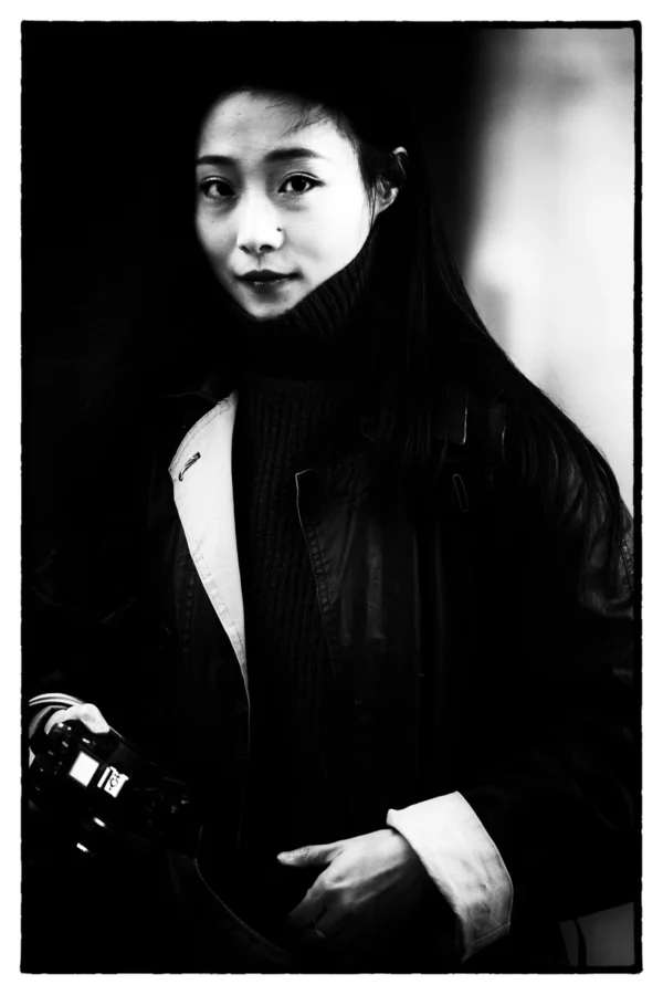 Thierry · 26/01/2026 · 2560 x 3840 Cette photographie en noir et blanc met en scène une femme comme sujet principal, au regard intense et légèrement penché. La composition est simple et centrée, avec un cadrage serré sur son visage et ses épaules. Elle porte un long manteau et tient un accessoire à la main.La lumière, douce et contrastée, éclaire son visage tout en créant des ombres marquées, accentuant les traits. L’ambiance est à la fois élégante et mystérieuse, renforcée par le style rétro et le flou d’arrière-plan. Les tons dominants sont les noirs profonds et les gris, typiques du noir et blanc.