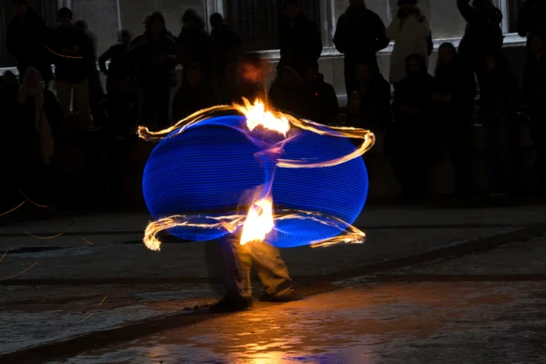 Ahmet · 24/01/2026 · 2560 x 1706 Cette photographie capture un artiste manipulant des boules de feu enflammées à l'intérieur de cerceaux lumineux bleus. Le sujet principal est le performeur, en mouvement dynamique, au centre de l'image.La composition est centrée, avec un cadrage serré sur l'action. Les couleurs dominantes sont les bleus des cerceaux et les teintes chaudes des flammes. La lumière est contrastée, avec des éclats vifs des feux et une ambiance nocturne en arrière-plan.Le style est artistique et spectaculaire, évoquant une ambiance théâtrale et captivante, typique des spectacles de rue.