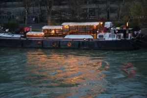 Laurence · 20/12/2025 · 3472 x 2320 Cette photographie montre un bateau-restaurant illuminé sur un plan d’eau, probablement un canal. Le sujet principal est ce bateau, décoré de guirlandes lumineuses chaudes.La composition est horizontale, avec une mise en avant du bateau au centre. Le cadrage est serré, capturant surtout l’eau et les lumières. Les couleurs dominantes sont les tons chauds (oranges, jaunes) des éclairages, contrastant avec le bleu nuit de l’eau.Le style est réaliste et légèrement nocturne, créant une ambiance chaleureuse et intimiste, évoquant une soirée paisible et élégante.