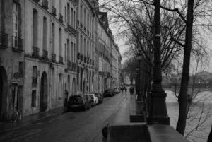 Laurence · 3472 x 2320 Cette photographie en noir et blanc capture une rue parisienne calme. Le sujet principal est l'alignement des bâtiments haussmanniens et le trottoir bordant un canal. La composition met en valeur une perspective linéaire, avec des voitures garées et des arbres dénudés.Le cadrage est serré, guidant le regard vers l’horizon. La lumière, douce et diffuse, crée des contrastes subtils. Les couleurs dominantes, bien que monochromes, jouent sur les nuances de gris.Le style est documentaire et intemporel, évoquant une ambiance mélancolique et paisible, typique des quartiers historiques parisiens.