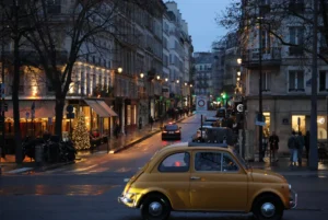 Laurence · 20/12/2025 · 3472 x 2320 Cette photographie met en scène une **voiture vintage jaune** (Fiat 500) comme sujet principal, au premier plan. La composition joue sur la perspective avec une **rue urbaine** légèrement en pente, bordée de bâtiments éclairés.Le **cadrage** est serré sur la voiture, mais inclut aussi des éléments architecturaux et des passants en arrière-plan. La **lumière** est douce, avec des éclairages chauds (ampoules, vitrines), créant une ambiance nocturne et intimiste. Les **couleurs dominantes** sont les tons jaunes, gris et bleus nocturnes.Le style évoque un **cliché romantique et nostalgique**, renforcé par l’éclairage tamisé et l’architecture classique. L’ambiance est **poétique et apaisante**, typique d’une ville européenne le soir.