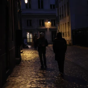 Laurence · 2320 x 2320 Cette photographie capture deux silhouettes marchant sur une rue pavée en pente, de nuit. Le sujet principal est ces deux personnes, dont les visages restent indistincts.La composition est simple et épurée, avec un cadrage serré sur les personnages et un arrière-plan de bâtiments éclairés par des lumières artificielles. La lumière, douce et tamisée, crée des ombres marquées et une ambiance mystérieuse.Les couleurs dominantes sont des teintes sombres et froides, avec des touches de jaune et d'orange provenant des éclairages. Le style est réaliste et sobre, évoquant une atmosphère nocturne calme et contemplative.