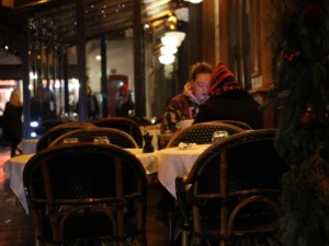 Laurence · 2773 x 2080 Cette photographie montre un restaurant élégant en soirée. Le sujet principal est un homme assis à une table, légèrement flou, suggérant une ambiance intimiste. La composition met en valeur des tables disposées en terrasse, avec des chaises en rotin et des nappes blanches.Le cadrage est serré sur les tables et l’espace immédiat autour. La lumière est douce et tamisée, avec des éclairages chauds provenant des lampes et des vitrines. Les couleurs dominantes sont les tons chauds (oranges, bruns) et les contrastes entre les lumières et les ombres.L’ambiance est chaleureuse et romantique, évoquant un lieu convivial et raffiné.