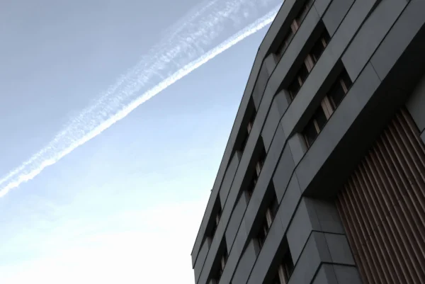Laurence · 3472 x 2320 Cette photographie met en scène un **immeuble moderne** en béton, sujet principal, avec une **structure géométrique anguleuse**. Le **cadrage serré** isole partiellement le bâtiment, accentuant ses lignes verticales.La **composition** joue sur le contraste entre l’architecture et le **sillage d’avion** traversant le ciel, ajoutant une touche dynamique. La **lumière** est naturelle, douce et légèrement voilée, créant une ambiance sereine.Les **couleurs dominantes** sont les tons **gris** (bâtiment) et **bleu pâle** (ciel), renforçant une atmosphère **minimaliste et contemporaine**. Le style est **épuré et photographique**, évoquant une **modernité urbaine**.