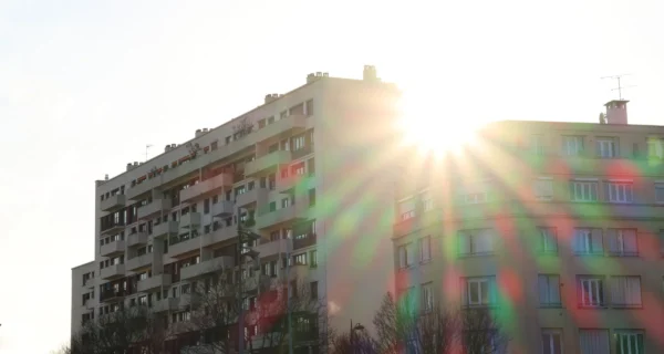 Laurence · 2679 x 1429 Cette photographie met en scène un immeuble résidentiel ancien, sujet principal, avec des balcons et des fenêtres.La composition est simple, centrée sur le bâtiment. Le cadrage est serré, capturant uniquement une partie du bâtiment et le ciel.La lumière, très forte, crée des effets de flare et des rayons solaires colorés (vert, rouge), dominant la scène. Les couleurs sont contrastées : gris et beige pour l’immeuble, avec des éclats de couleurs vives dus à la lumière.Le style est réaliste et brut, évoquant une ambiance à la fois urbaine et mélancolique, renforcée par les dégradations visibles sur le bâtiment.