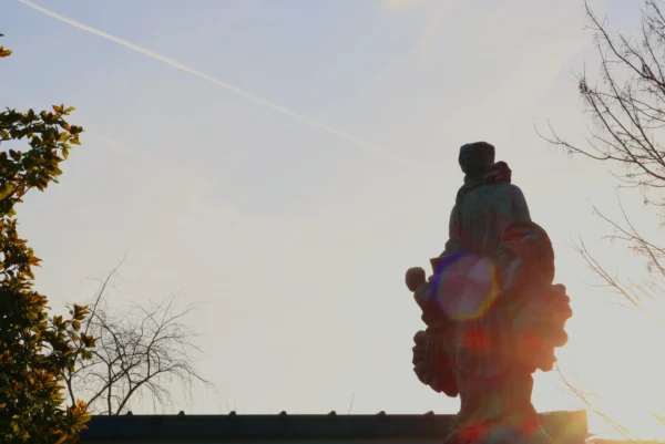 Laurence · 3472 x 2320 Cette photographie met en scène une statue silhouettée, probablement religieuse, vue de dos. La composition joue sur le cadrage serré et la lumière rasante du soleil couchant, créant un effet de contre-jour qui accentue les contours et génère une auréole lumineuse autour du sujet.Les couleurs dominantes sont les tons chauds (oranges, jaunes) contrastant avec les teintes froides du ciel. Le style est épuré et dramatique, renforcé par les branches nues en arrière-plan. L’ambiance est à la fois mystique et contemplative, évoquant sérénité et spiritualité.