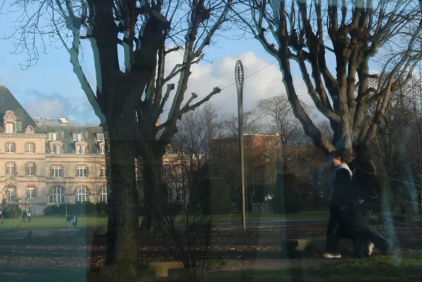 Laurence · 3472 x 2320 Cette photographie montre un parc en hiver avec un **sujet principal flou** : une personne en mouvement, vêtue d’un manteau sombre. La **composition** est équilibrée avec des arbres nus en premier plan et un bâtiment classique en arrière-plan. Le **cadrage** est légèrement bas, encadré par les branches.La **lumière** est douce, avec des ombres marquées, suggérant un ensoleillement hivernal. Les **couleurs dominantes** sont les tons froids : gris, brun et bleu pâle. Le style est **réaliste et naturel**, avec une ambiance **calme et mélancolique**, renforcée par la saison et la lumière tamisée.