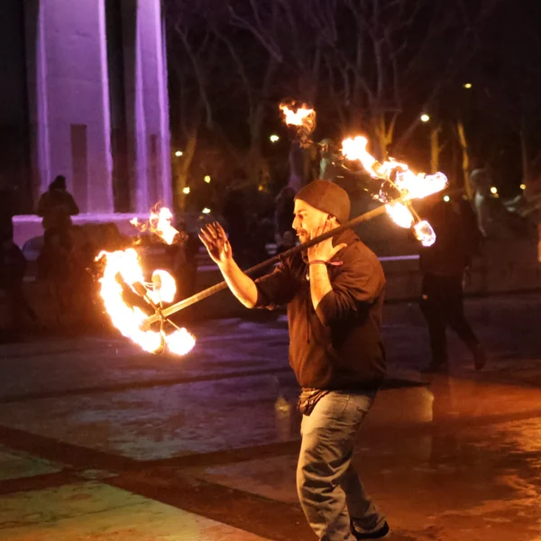 Laurence · 2157 x 2157 Cette photographie capture un artiste de feu comme sujet principal. Il manipule des torches enflammées avec des bâtons, au centre du cadre.La composition est dynamique, avec une mise au point sur le performer. Le cadrage est serré, mettant en valeur son action. Les flammes éclairent la scène, créant un contraste saisissant avec l'obscurité environnante.Les couleurs dominantes sont les teintes chaudes des flammes (orange, jaune) et les tons sombres de la nuit. Le style est réaliste et captivant, évoquant une ambiance spectaculaire et intense, presque mystique.