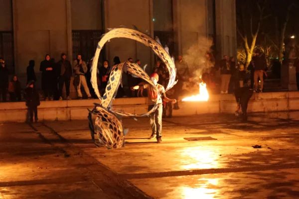 Laurence · 2811 x 1874 Cette photographie capture un artiste manipulant des objets enflammés, probablement des massues à feu, lors d’une performance nocturne. Le sujet principal est l’artiste en mouvement, entouré de flammes vives.La composition est dynamique, avec un cadrage serré sur l’action. L’éclairage est dramatique, mettant en valeur les flammes et créant des contrastes forts entre lumière et obscurité. Les couleurs dominantes sont les teintes chaudes des flammes (orange, jaune) et les tons sombres de la nuit.L’ambiance est intense et spectaculaire, évoquant l’énergie et le danger du spectacle. Le style relève du street performance artistique.
