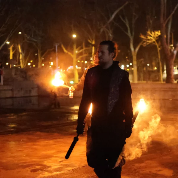Laurence · 2320 x 2320 Cette photographie met en scène un homme comme sujet principal, vêtu d’un long manteau sombre, marchant avec des torches allumées dans chaque main. La composition est dynamique, centrée sur le personnage en mouvement. Le cadrage est serré, isolant le sujet sur un fond flou de nuit urbaine.Les flammes éclairent la scène, créant une lumière chaude et contrastée. Les couleurs dominantes sont les tons chauds (oranges, rouges) des torches, contrastant avec le noir du manteau et l’obscurité ambiante. Le style est dramatique et cinématographique, évoquant une ambiance mystérieuse et intense, presque théâtrale.