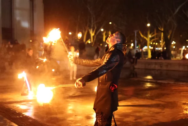 Laurence · 3472 x 2320 Cette photographie capture un individu en train de manipuler des torches enflammées. Le sujet principal est au centre, vêtu d’une veste sombre, les bras écartés, avec des flammes vives autour de lui.La composition est dynamique, avec un cadrage serré qui met en valeur l’action. L’ambiance est nocturne, éclairée par les flammes et quelques lumières artificielles en arrière-plan. Les couleurs dominantes sont les tons chauds (oranges, jaunes) des feux, contrastant avec les teintes sombres de la nuit.Le style évoque un spectacle ou une tradition, créant une ambiance intense et mystérieuse.