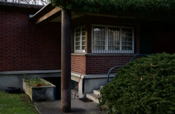 Paul C. · 17/01/2026 · 5373 x 3503 Cette photographie montre une maison en briques avec un style architectural sobre et moderne. Le sujet principal est la façade partielle de la maison, incluant une fenêtre à carreaux et une porte latérale.La composition est simple, avec un cadrage serré sur la structure et un jardin en arrière-plan. La lumière semble naturelle, mais un peu tamisée, créant des ombres douces. Les couleurs dominantes sont les tons bruns des briques et verts des plantes. L’ambiance est calme et résidentielle, évoquant une demeure tranquille et bien entretenue.