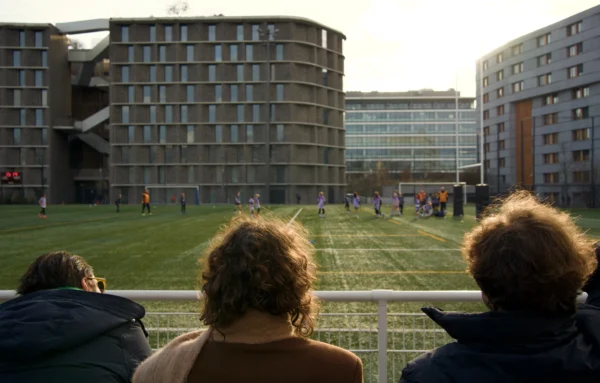 Paul C. · 17/01/2026 · 5910 x 3776 Cette photographie montre un groupe de spectateurs regardant un match de football depuis une tribune. Le **sujet principal** est le terrain de jeu et les joueurs en action, encadrés par des bâtiments modernes en arrière-plan.La **composition** est dynamique, avec une perspective légèrement basse. Le **cadrage** met en valeur les spectateurs au premier plan, créant un effet de profondeur. La **lumière** semble naturelle, probablement en journée, avec des ombres douces. Les **couleurs dominantes** sont des tons neutres (beige, gris) et verts (pelouse), adoucis par des touches de rouge et bleu dans les vêtements.Le **style** est réaliste et documentaire, évoquant une ambiance **quotidienne et sportive**, presque intimiste malgré la présence de nombreux spectateurs.
