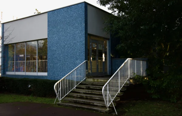 Paul C. · 17/01/2026 · 5877 x 3758 Cette photographie montre un bâtiment moderne aux tons pastel, principalement bleu clair. Le sujet principal est l'entrée accessible par un escalier métallique avec une rampe. La composition est symétrique, avec des lignes droites et une structure épurée.Le cadrage est serré sur l'entrée, encadrée par la végétation. La lumière semble naturelle, douce et légèrement tamisée, créant des ombres douces. Les couleurs dominantes sont le bleu, le gris et le vert des plantes. Le style architectural est contemporain et minimaliste, évoquant une ambiance calme et fonctionnelle.