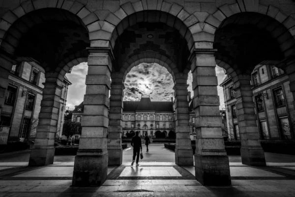 Marc · 17/01/2026 · 5781 x 3854 Cette photographie en noir et blanc met en scène une **architecture classique** avec des **arcades imposantes** en pierre. Le **sujet principal** est l’ouverture centrale, encadrée par les arches, qui donne sur une place urbaine.La **composition** est symétrique, avec des lignes directrices convergentes vers le centre. Le **cadrage** est serré, accentuant la profondeur et la perspective. La **lumière**, contrastée, crée des ombres marquées, renforçant le côté dramatique. Les **couleurs dominantes** sont les nuances de gris, typiques du noir et blanc.Le **style** est **sobre et architectural**, évoquant une **ambiance solennelle et intemporelle**.