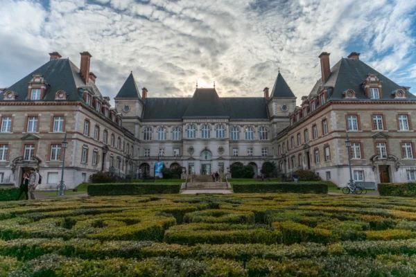 Marc · 17/01/2026 · 7008 x 4672 Cette photographie met en scène un **château majestueux** aux allures classiques, avec une façade symétrique et des toits en ardoise. Le **sujet principal** est ce bâtiment imposant, entouré d’un **jardin géométrique** bien entretenu.La **composition** est équilibrée, avec un **cadrage centré** sur l’entrée principale. La **lumière**, douce et tamisée, suggère un coucher de soleil ou un temps nuageux, créant des contrastes subtils. Les **couleurs dominantes** sont les tons **beige, gris et verts**, adoucis par des touches de bleu dans le ciel.Le **style** est **classique et historique**, évoquant une ambiance **sereine et intemporelle**, presque mystérieuse.