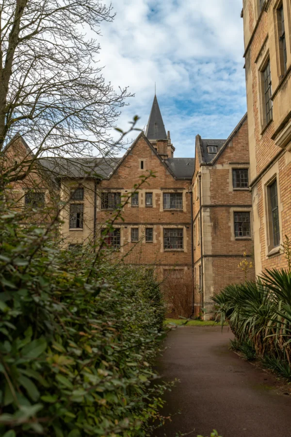 Marc · 17/01/2026 · 4672 x 7008 Cette photographie met en scène un **cour intérieure d’un bâtiment historique**, probablement une université ou un collège. Le **sujet principal** est l’architecture gothique, avec ses murs en pierre, ses fenêtres à meneaux et sa tour crénelée.La **composition** est symétrique, avec un chemin central bordé de végétation. Le **cadrage** est serré, guidant le regard vers le fond. La **lumière** naturelle, douce et légèrement voilée, crée des ombres douces. Les **couleurs dominantes** sont les tons beiges et bruns des pierres, contrastés par le vert des plantes et le bleu pâle du ciel.Le style est **classique et sobre**, évoquant une ambiance **sereine et intemporelle**.