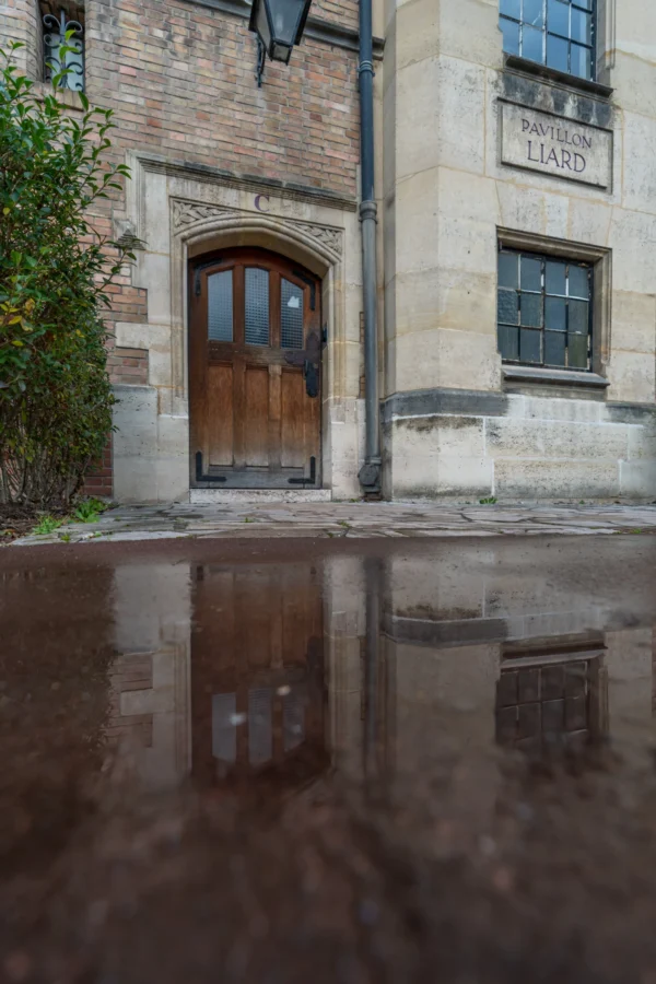 Marc · 17/01/2026 · 4387 x 6580 Cette photographie met en scène une entrée d’un bâtiment ancien en pierre, avec une porte en bois et des fenêtres étroites. La composition joue sur la symétrie et la réflexion dans une flaque d’eau au premier plan, créant un effet miroir.Le cadrage est serré, centré sur l’architecture. La lumière est douce, naturelle, avec des ombres marquées, suggérant un moment calme, probablement en journée. Les couleurs dominantes sont les tons beiges et gris de la pierre, adoucis par les reflets sombres de l’eau.Le style est sobre et minimaliste, évoquant une ambiance historique et contemplative.