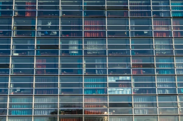 Marc · 17/01/2026 · 3581 x 2372 Cette photographie met en scène la façade d’un immeuble moderne aux multiples fenêtres. Le sujet principal est la répétition géométrique des vitres reflétant des éléments urbains et des couleurs variées.La composition repose sur un cadrage serré, mettant en valeur les lignes verticales et horizontales des baies vitrées. La lumière, naturelle et diffuse, accentue les reflets et les contrastes entre les teintes.Les couleurs dominantes sont des bleus, gris et touches de rouge et vert, créant une ambiance à la fois froide et dynamique. Le style est épuré et architectural, évoquant une ville contemporaine et urbaine.
