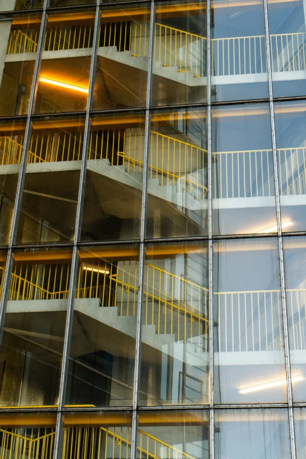 Marc · 17/01/2026 · 3440 x 5160 Cette photographie met en scène un bâtiment moderne avec une façade en verre réfléchissant. Le sujet principal est une série d'escaliers extérieurs métalliques, partiellement visibles à travers les vitres. La composition joue sur les reflets et les lignes verticales, créant une impression de profondeur et de symétrie. Le cadrage est serré, mettant en valeur les détails architecturaux.La lumière naturelle, douce et légèrement dorée, accentue les contours et les transparences. Les couleurs dominantes sont les teintes grises et bleutées, avec des reflets dorés et blancs. Le style est épuré et contemporain, évoquant une ambiance urbaine et sophistiquée.
