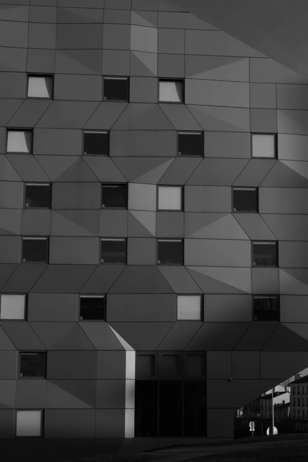 Don · 3275 x 4912 Cette photographie met en scène un bâtiment moderne aux façades géométriques. Le sujet principal est l’architecture, marquée par des panneaux triangulaires et des fenêtres rectangulaires alignées. La composition est épurée, avec un cadrage serré sur la structure.La lumière, naturelle et contrastée, accentue les ombres et les jeux de volumes. Les couleurs dominantes sont les gris et noirs, renforçant une ambiance minimaliste et contemporaine. Le style est architectural et photographique, avec un rendu noir et blanc qui souligne la texture et la rigueur des formes. L’ambiance est à la fois froide et élégante, évoquant une modernité urbaine.