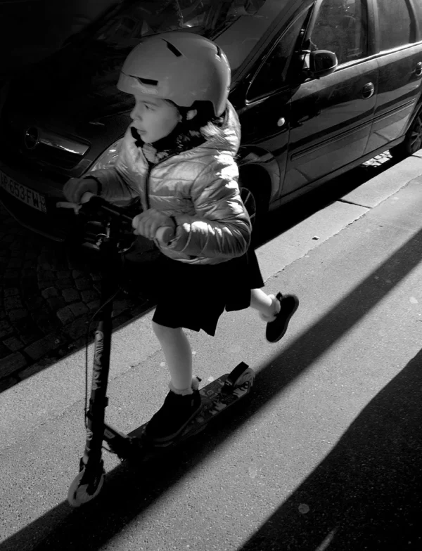 Pascale · 24/01/2026 · 765 x 1000 Cette photographie met en scène un enfant en train de monter sur un scooter, vêtu d'une veste épaisse et d'un casque. Le sujet principal est bien cadré, occupant une grande partie de l'image.La composition est dynamique, avec un angle bas qui accentue la perspective. La lumière, naturelle et contrastée, crée des ombres marquées, renforçant l'ambiance dramatique. En noir et blanc, les tons dominants varient du blanc à des gris profonds, soulignant les textures et les formes.Le style est documentaire et spontané, évoquant une ambiance urbaine et un moment de quotidien capturé avec authenticité.