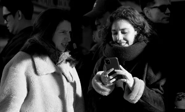 Pascale · 24/01/2026 · 966 x 588 Cette photographie en noir et blanc met en scène deux personnes comme sujets principaux. La composition montre une interaction sociale, l’une tenant un téléphone tandis que l’autre semble observer ou discuter.Le cadrage est serré, centré sur les visages et les épaules. La lumière est tamisée, créant des contrastes doux et des ombres légères, typiques d’un environnement nocturne ou peu éclairé.Le style est documentaire et spontané, évoquant une ambiance authentique et intime. Les tons dominants, en niveaux de gris, renforcent une atmosphère à la fois moderne et nostalgique.