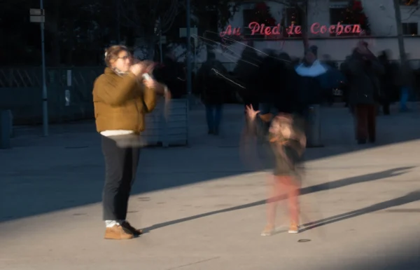Alice · 03/01/2026 · 3429 x 2200 Cette photographie capture une scène de rue où le sujet principal est une femme et une enfant jouant avec des baguettes, probablement lors d’un spectacle de rue. La composition est dynamique, avec un cadrage serré sur les deux personnages en mouvement. L’arrière-plan est flou, mettant en valeur les sujets.La lumière est naturelle, douce, avec des ombres peu marquées. Les couleurs dominantes sont les tons chauds : orange du vêtement de l’enfant, jaune du haut de la femme, et des teintes neutres pour le reste. Le style est documentaire et spontané, évoquant une ambiance joyeuse et artistique dans un cadre urbain.